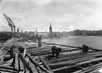 Svart-vit bild från hög höjd med utsikt mot staden och Mälaren. I bilden syns fyra män lutade ner över sitt arbete med timret. I bakgrunden till vänster syns en lyftkran.