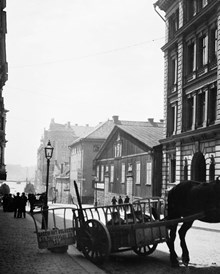 En hästkärra med mjölkkrukor står vid Grevgatan 5 (nu 15). Skylten på kärran hänvisar till S:t Paulsgatan 22. Efter trähuset t.h. ligger hörnet av Riddargatan. I fonden söderut skymtar Nybroviken