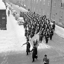 Svea Livgarde på Riddargatan i hörnet av Artillerigatan på väg till Riksdagens högtidliga öppnande. Uniformerna är av 1820-tals modell. På fanan markeras fältslagen