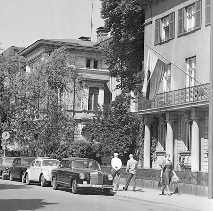 Två män och en kvinna syns på trottoaren. En flagga hänger från en fasad. Bilar står parkerade längs gatan.