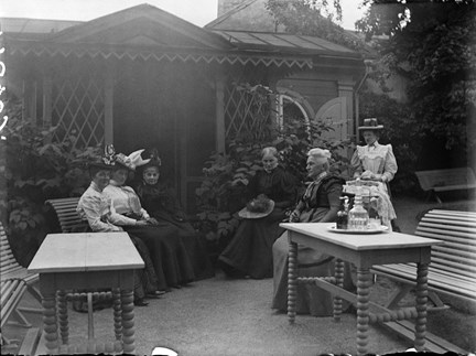 Ett lusthus med en veranda, täckt av växtlighet. Framför sitter sex stycken kvinnor i hattar och långa kjolar och blusar på trädgårdssoffor. En bricka med dricka och glas står på det ena bordet. 