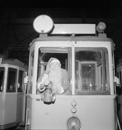 Jultomten tittar ut ifrån ett fönster i förarhytten på en spårvagn och håller en lyckta i handen. 