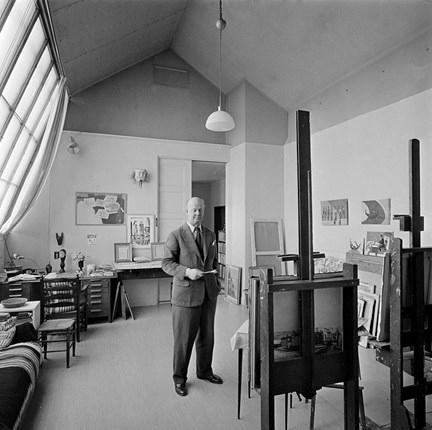 Södermalmstorg 4. Kurt Jungstedt i sin ateljé. Konstnären står framför ett staffli med en pensel i handen. Ljuset kommer från det stora ateljéfönstret.