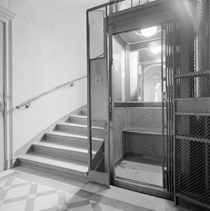 Trapphus. En trappa leder uppåt, till höger om den syns en stilren hiss i avskalad design. Golvet framför hissen är rutmönstrat, förmodligen av mönsterlagd marmor.