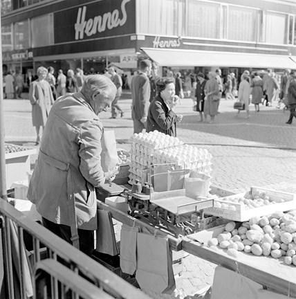 På ett torg står en äldre man och säljer potatis och ägg. En kvinna går förbi och stoppar precis en brödbit i munnen. I bakgrunden syns en butiksbyggnad med stora skyltfönster. 