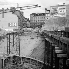 Mäster Samuelsgatans bro österut mot Malmskillnadsgatan. Tunnelbanebyggandet pågår under blivande Sveavägen. T.v. Mäster Samuelsgatan 40, i mitten Malmskillnadsgatan 40. Reklam på kv. Hästryggen