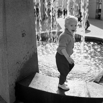 Ett barn i 3-4-årsåldern går på kanten till en fontän med rund liten bassäng. Solen blänker i vattnet. Barnet skrattar. 