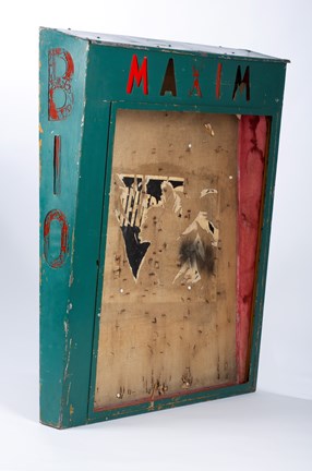 Ett skåp fotat snett från vänster med mörkgrön ram och texten "Maxim" i överkant. Till vänster om ramen står texten: "Bio."