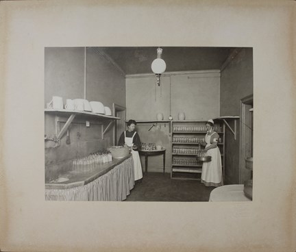 Två kvinnor i sjuksköterskedräkt och med vitt förkläde i ett rum med hyllor