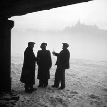 Tre män står under Järnvägsbron i Gamla Stan och delar på en butelj. Laurinska huset i fonden