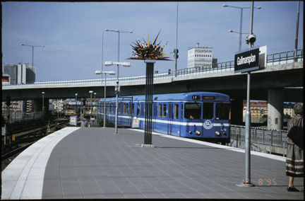 Ett blått tunnelbanetåg rullar in till Gullmarsplans tunnelbanestation år 1995. Mitt i bild, på perrongen står Leif Tjerneds målade stålskulptur. 