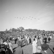 En folksamling på Ladugårdsgärdet ser på flyguppvisning. I bakgrunden skymtar Borgen