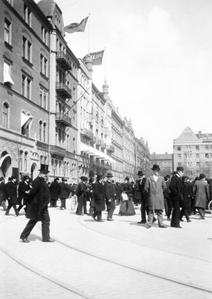 Till vänster i bild syns fasader av byggnader med fem eller sex våningar. Flaggor vajar på taket och nere på gatan syns ett femtiotal män och kvinnor.