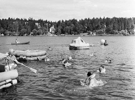 Människor som badar i en sjö. I vattnet finns också båtar