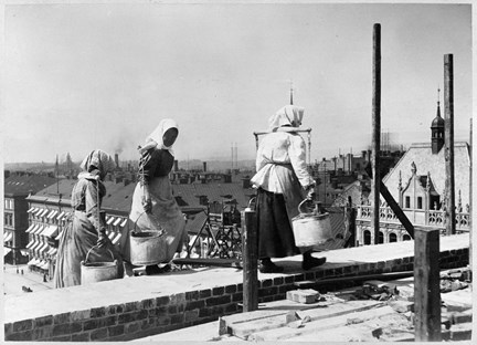 Svart-vit bild med tre kvinnor på toppen av ett tak. I bakgrunden hustak och skorstenar. Kvinnorna är klädda i kjol, skjorta och sjal över huvudet. De bär på hinkar i ok över axlarna. 