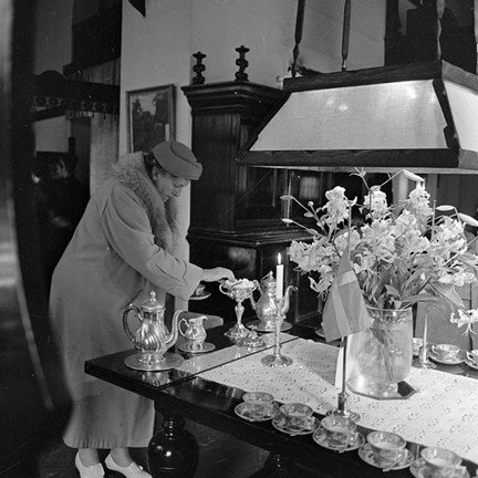 En äldre kvinna i pälskantad kappa och hatt tar för sig kaffe från ett bord med spetsduk och uppdukade kaffekoppar. Hon tar just en bit socker.