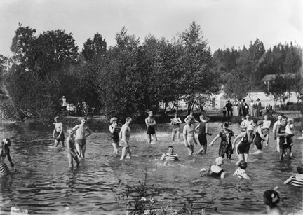 En mängd män, kvinnor och barn i olika åldrar badar intill en strand. I bakgrunden syns träd och ett äldre trähus. Män och pojkar har badbyxor, kvinnorna baddräkter med korta ärmar och ben.