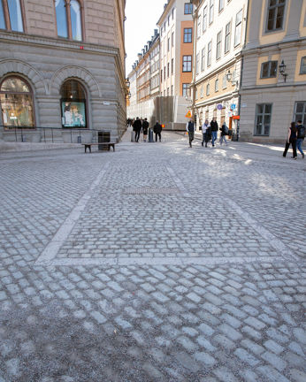 Foto taget på dagtid av ett torg och en gata i bakgrunden. Torgets stenläggning med L-form syns tydligt
