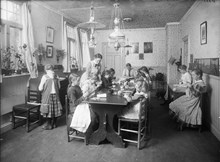 Interiör från Hagalunds arbetsstuga med flickor och kvinnor som sitter vid bord och läser, syr och stickar.