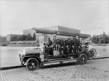 Brandbil, troligen en Scania Vabis, med brandmän  på Södra Blasieholmskajen.  I bakgrunden Slottet. Troligen är det brandkårens första motorspruta som fotograferas 30 juli 1918.