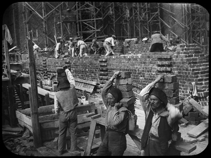 Svart-vit bild. Två män i förgrunden bär tegelstenar på ryggen och en tredje man förbereder sitt lyft. I bakgrunden syns murade väggar, byggställningar och murare som arbetar.