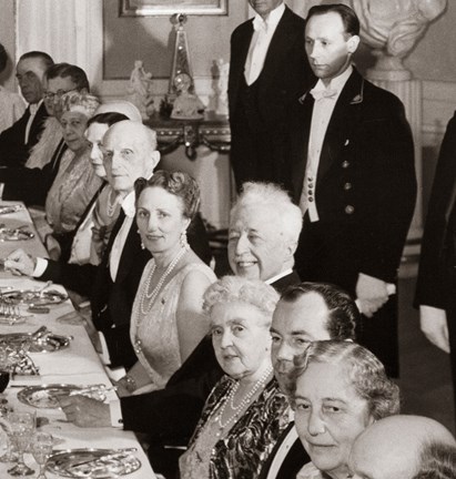 Svartvitt fotografi från middagsbjudning i matsalen på Waldemarsudde. Gästerna på ena långsidan av bordet har vänt huvudet mot kameran, alla är finklädda och prinsen ler stort. I bakgrunden står personalen uppradade. 