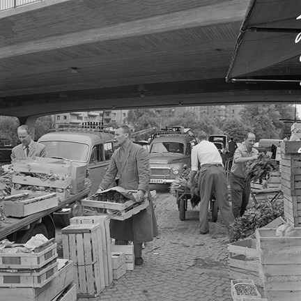 Under en låg bro står bilar och små lastbilar parkerade. Män bär och staplar trälådor med grönsaker.