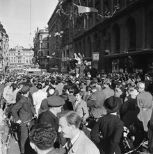 Fredsdagen den 7.e maj 1945 på Kungsgatan i Stockholm. 
Glada folkmassor längs gatan och från husfasadernas fönster kastar människor papper och girlanger.