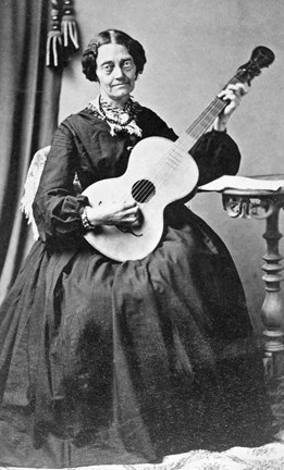 Porträttfotografi på en kvinna i femtio till sextioårsåldern. Kvinnan bär en klänning från andra hälften av 1800-talet och slår an ett ackord på en gitarr. På ett litet pelarbord intill ligger det ett papper, möjligen noter.