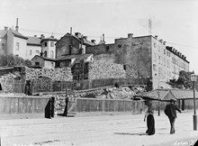 En kvinna och en man passerar ett stort vedlager vid Birger Jarlsgatan 32-30, kv. Häckelfjäll Större. Innan 1885 hette gatan Stora Träskgatan. Nuvarande Birger Jarlsgatan 34-32, kv. Sparbössan