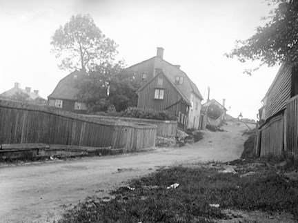 En slingrande väg utan stenbeläggning leder svagt uppåt. På Vänster sida ser vi flera små hus av trä eller sten. Till höger ett plank. Inga människor syns i bilden.