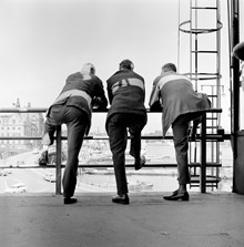 Slussen, utsikt mot Gamla Stan och Skeppsbron. Tre män står lutade mot ett räcke. Till höger en brandstege