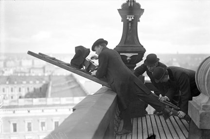 Högt upp på kanten till ett torn, lutar sig en filmare mot en stege som ligger över kanten. Två män håller i stegen medan filmaren matar sin kamera med film. I bakgrunden syns taket till ett slott.