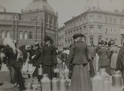 Kvinnor rör sig på torget. En rad mjölkkannor i förgrunden.
