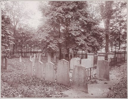 I den svart-vita bildens nedre del syns ett tjugotal gravstenar omgivna av ett staket, i den övre delen syns grönskande träd.