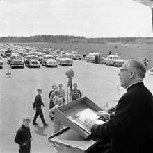Skarpnäcks flygfält. Biskop Odd Hagen predikar på Sveriges första drive-in-gudstjänst. Tusen bilar stod tätt uppställda och ca sextusen personer deltog. Altaret var rest på ett lastbilsflak.