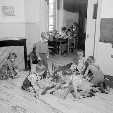 Barn leker med träleksaker på golvet. I bakgrunden sitter tre barn och en kvinna vid ett bord. 