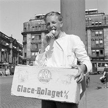 Tegelbacken. En ung glassförsäljare äter glass, med en glasslåda från Glace-Bolaget (GB) på magen. Centralpalatset i bakgrunden