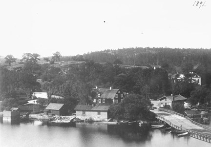 Liljeholmsbron till höger i bild, äldre bebyggelse ligger i grönska. Några småbåtar ligger förtöjda vid bron och bryggor.
