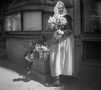 En äldre kvinna i kjol, förkläde, blus och huckle står på en gata och säljer blommor. Bredvid henne på en korg står kärl med blommor och buketter. 