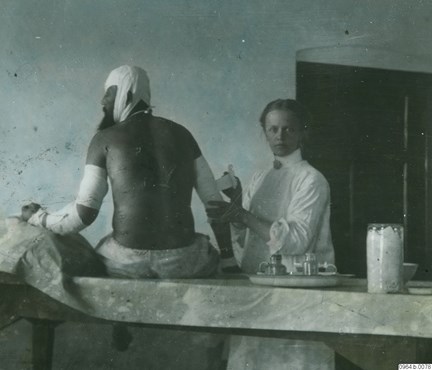 Kolorerat fotografi som visar en man, med ryggen mot kameran, och en vitklädd kvinna som förbinder hans arm.