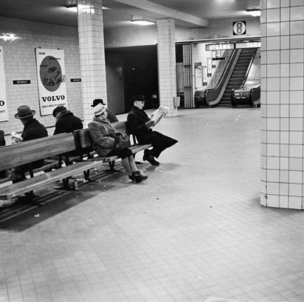 En tunnelbanestation under jord där väggar och pelare är klädda med ljust kakel. På bänkar sitter väntande passagerare, bland annat en man som läser tidningen. I fonden en rulltrappa. På väggarna syns stora reklamtavlor.