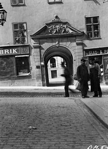 Några män står i hörnet av Mariagränd och Götgatan. I bakgrunden ligger portalen till Götgatan 16, Ebba Brahes palats