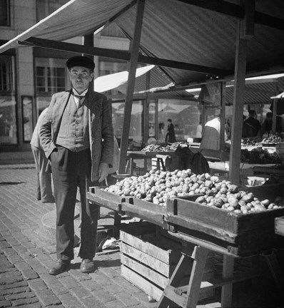 Vid ett torgstånd med flera lårar fulla med potatis står en man, försäljaren. Han bär en väst under kavajen och en keps på huvudet och har den ena handen i fickan. Solen skiner.