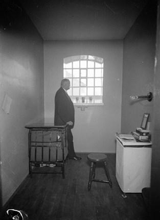 En man i kostym står vid fönstret i en kal, sparsamt möblerad fängelsecell.