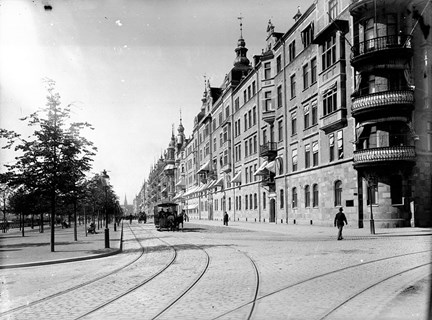 I marken på en bred boulevardliknande gata ligger spårvägsspår. Till höger i bild syns påkostade flerfamiljshus i sen 1800-talsstil. I fonden kommer en hästdragen spårvagn.