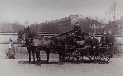 Två förspända hästar, sex brandmän på vagnen. En kvinna med hatt och hopfällt parasoll passerar.