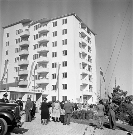 Människor står samlade nedanför ett högt, nybyggt vitt hus i sju våningar med balkonger. Vid husets entré är en skylt uppsatt med texten "Lägenheter som växer". 
