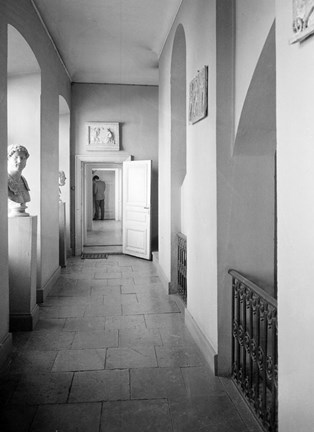 En korridor kmed stengolv. Rakt fram en dörr, längst bort står en man. Till vänsterfönsternischer med byster/statyer.