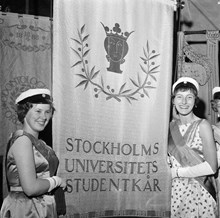 Hantverkargatan 1. Stockholms högskola blev Stockholms universitet. Invigning av det nya universitetet firas i Stadshuset. Två flickor i studentmössa och långklänningar står vid studentkårens fana